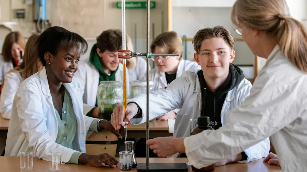 Lärare instruerar två elever i ett kemiskt experiment. I bakgrunden andra elever. Alla har vita rockar.